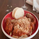 A vertical shot of a red bowl filled with penne pasta and thick Sicilian Red Gravy, topped with two large meatballs and a dusting of Pecorino Romano cheese, served with two slices of bread and a glass of red wine in the background.