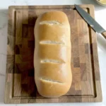 Homemade golden-brown French bread loaf sitting on a wooden cutting board with a butter knife and a small dish of butter on a white marble countertop.